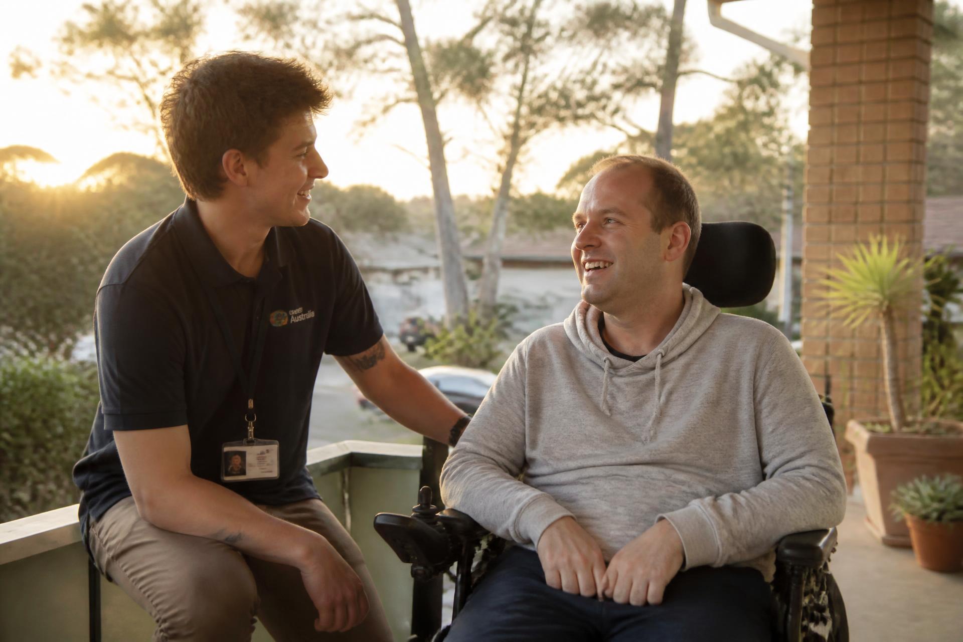 A support worker and a man using a wheelchair sharing a laugh on a sunlit veranda at sunrise