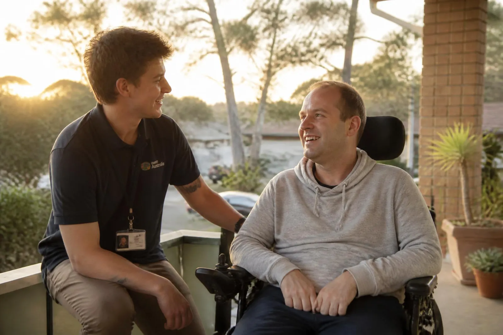 A support worker and a man using a wheelchair sharing a laugh on a sunlit veranda at sunrise